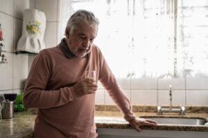 An older man stands in the kitchen holding a glass of water and considering how a heart attack can lead to depression.